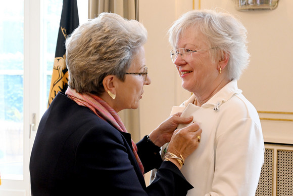 Zwei ältere Frauen, eine steckt der anderen eine Anstecknadel an, beide lächeln.