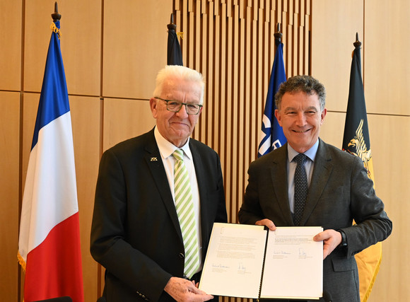 Ministerpräsident Winfried Kretschmann (links) und Franck Leroy (rechts), Präsident der Region Grand Est, stehen vor Fahnen und haben die unterzeichneten Positionspapiere in der Hand.