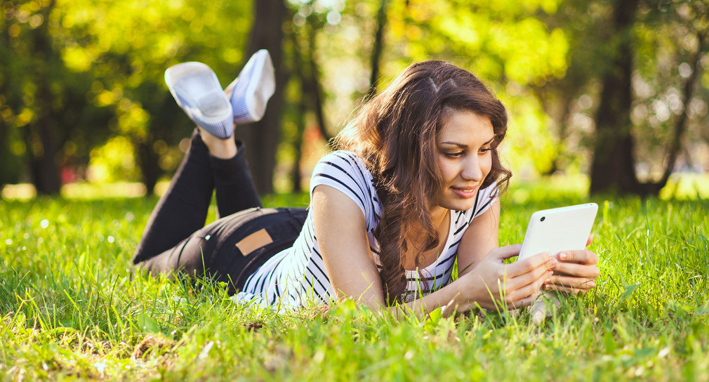 Eine Frau liegt mit einem Tablet in der Hand auf einer Wiese in einem Park.