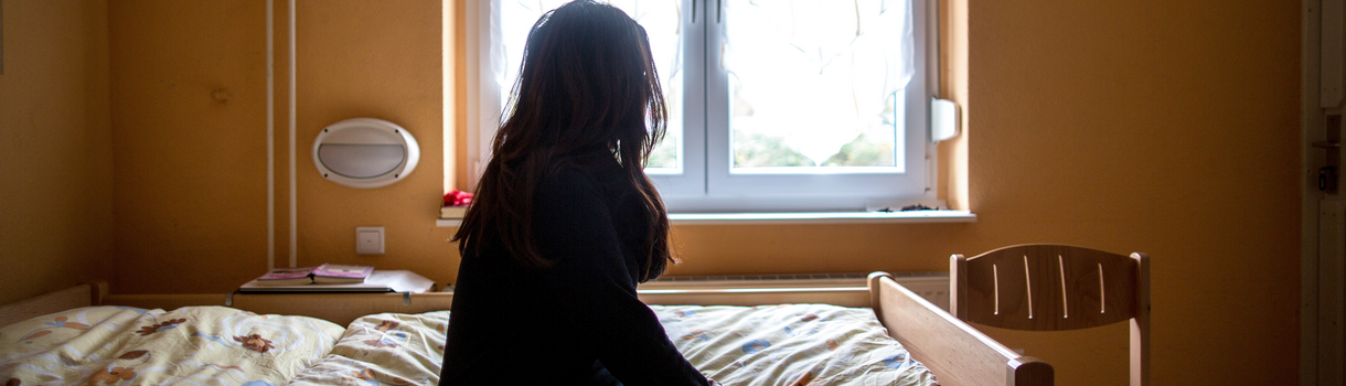 Eine Frau sitzt zum Fenster blickend in einem Frauenhaus auf einem Bett.