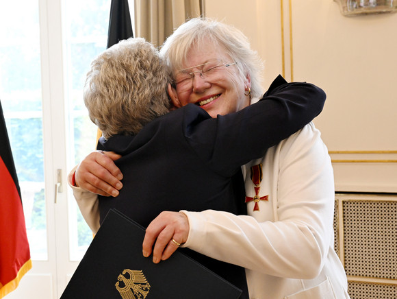Zwei ältere Frauen umarmen sich herzlich, eine hält eine Mappe mit dem deutschen Bundesadler, im Hintergrund eine deutsche Flagge.