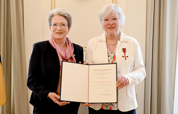 Zwei ältere Frauen, beide halten eine Mappe mit einer Urkunde, die andere trägt eine Auszeichnung am Revers.