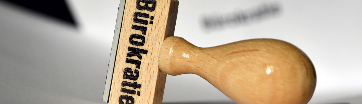 Ein Stempel mit der Aufschrift "Bürokratie" liegt auf Papieren.