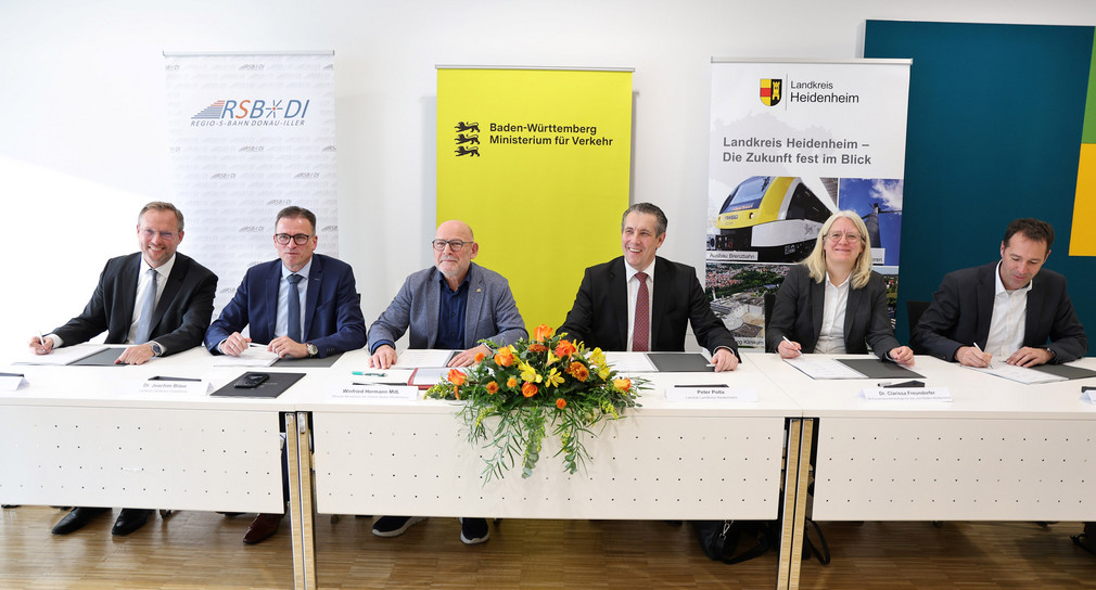 Eine Gruppe von Menschen sitzt am Tisch und schaut in die Kamera. Vor ihnen liegen Dokumente zum unterschreiben.