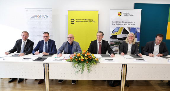 Eine Gruppe von Menschen sitzt am Tisch und schaut in die Kamera. Vor ihnen liegen Dokumente zum unterschreiben. Eine Gruppe von Menschen sitzt am Tisch und schaut in die Kamera. Vor ihnen liegen Dokumente zum unterschreiben.