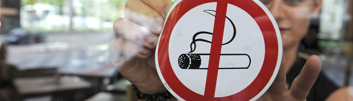 Eine Frau befestigt einen „Rauchen-Verboten“-Aufkleber an einer Scheibe (Bild: © dpa).