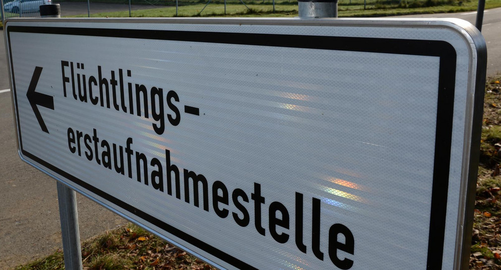 Ein Schild mit der Aufschrift „Flüchtlingserstaufnahmestelle“, im Hintergrund ein Fahrzeug an einer Einfahrt.
