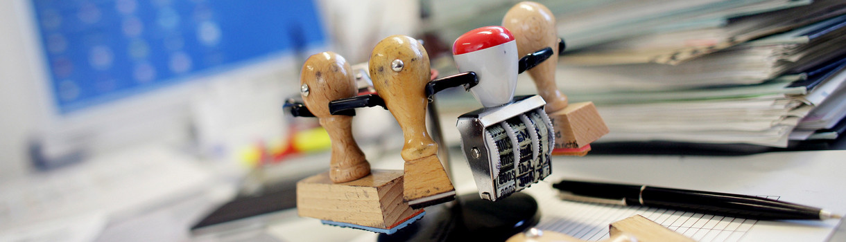 Symbolbild: Ein Stempelkarussell (Stempelhalter) steht neben einem Stapel Akten auf einem Schreibtisch in einer Behörde. (Bild: dpa)