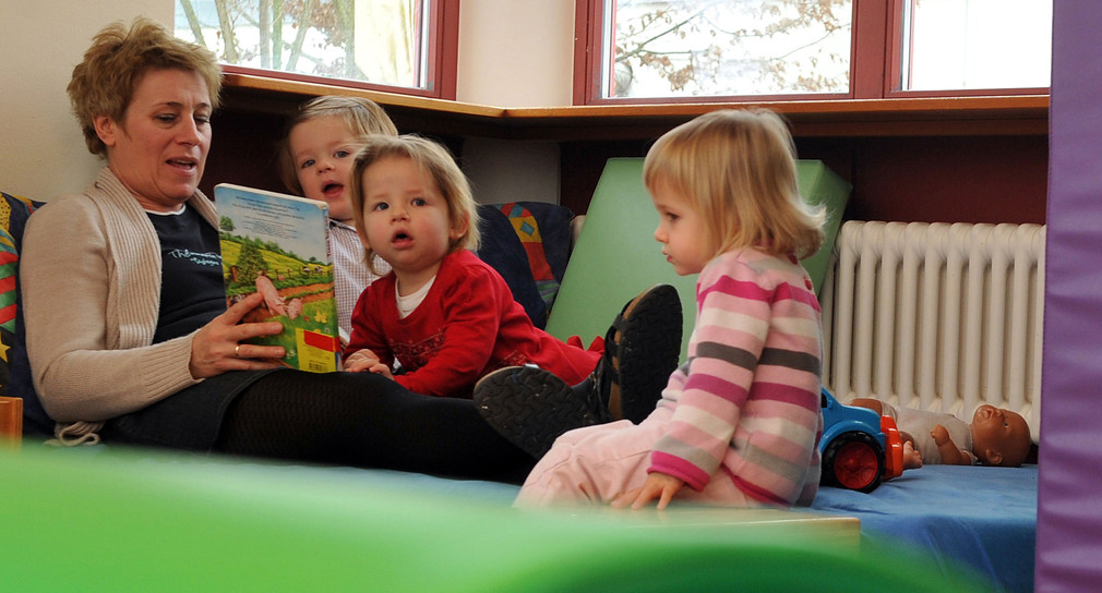 Eine Erzieherin ließt drei Kleinkindern aus einem Buch vor (Bild: © dpa).