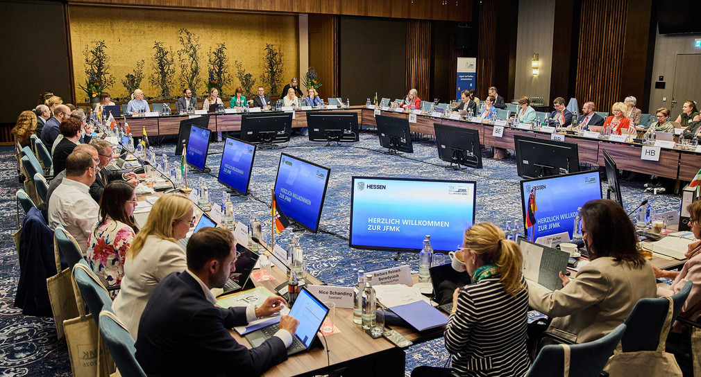 Teilnehmende der Jugend- und Familienministerkonferenz 2026 sitzen mit Blick auf mehrere kreisförmig angeordnete Bildschirme an einem großen Konferenztisch.
