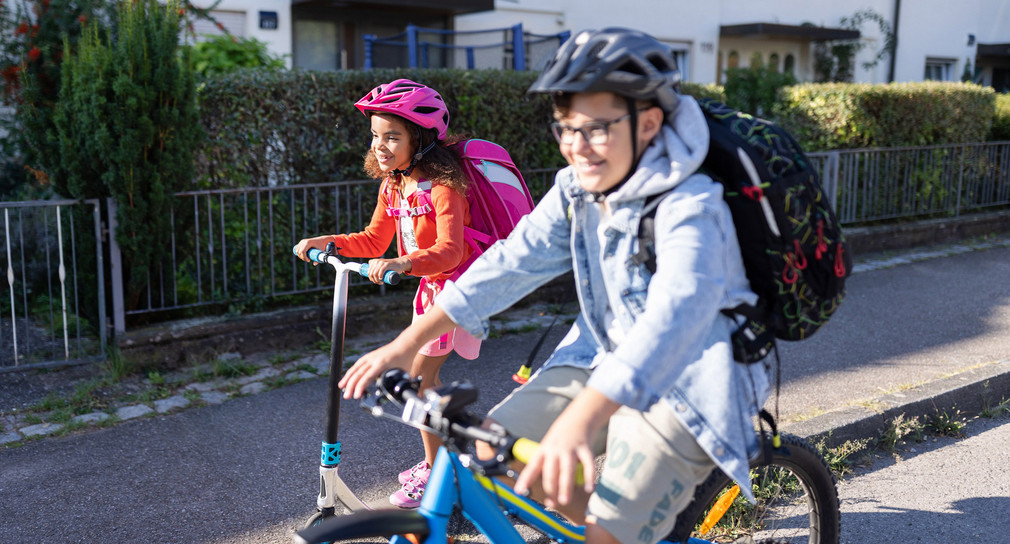 Zwei Kinder fahren zur Schule. Der Junge mit einem Fahrrad, das Mädchen mit einem Roller. Beide Kinder tragen einen Helm.