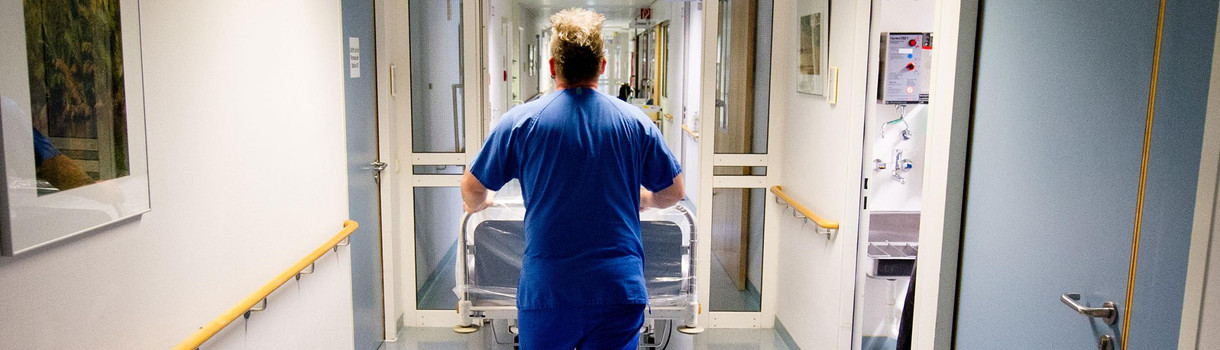 Ein Krankenpfleger schiebt in einer Klinik ein Krankenbett über einen Flur. (Foto: © dpa)