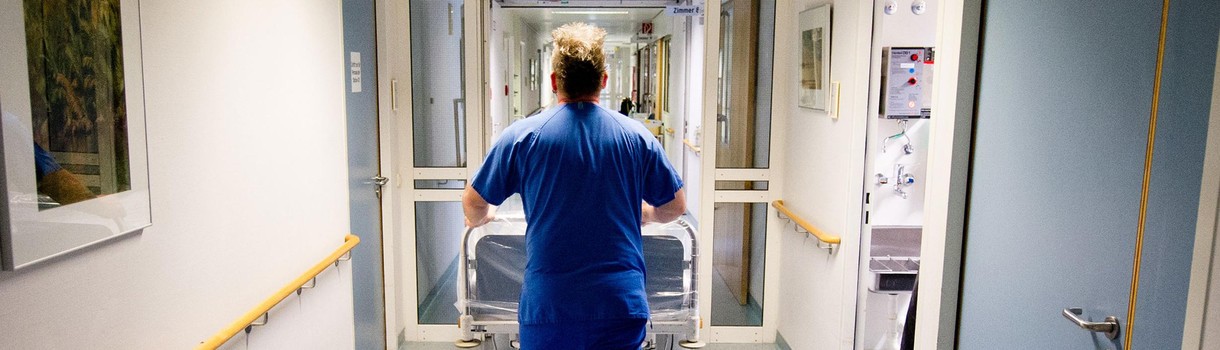 Ein Krankenpfleger schiebt in einer Klinik ein Krankenbett über einen Flur. (Foto: © dpa)