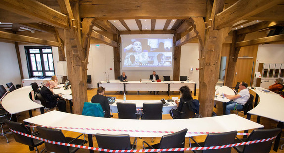 Eine Gemeinderatssitzung in einem Sitzungssaal mit zugeschalteten Mitgliedern auf einer Videoleinwand.