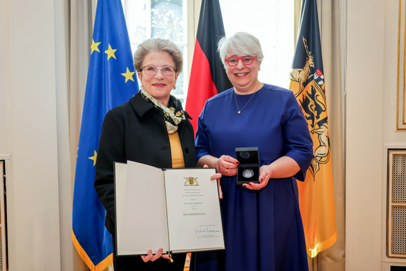 Staatsrätin Barbara Bosch (links) überreicht Irene Armbruster (rechts) die Staufermedaille und eine Urkunde. Im Hintergrdund stehen Fahnen.