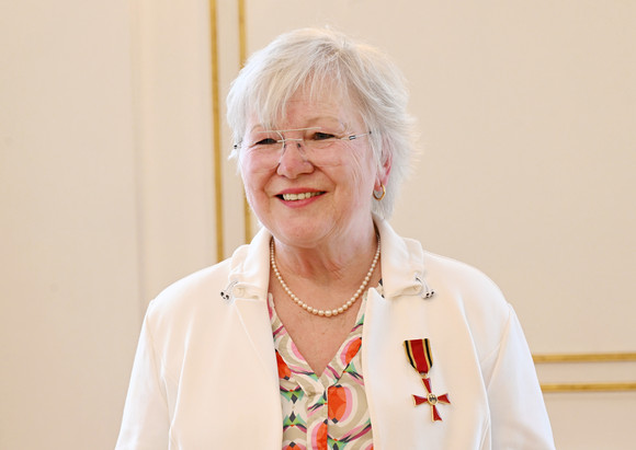 Ältere Frau mit weißem Haar, Brille und Perlenkette, trägt eine weiße Jacke mit einem Orden am Revers, und lächelt.