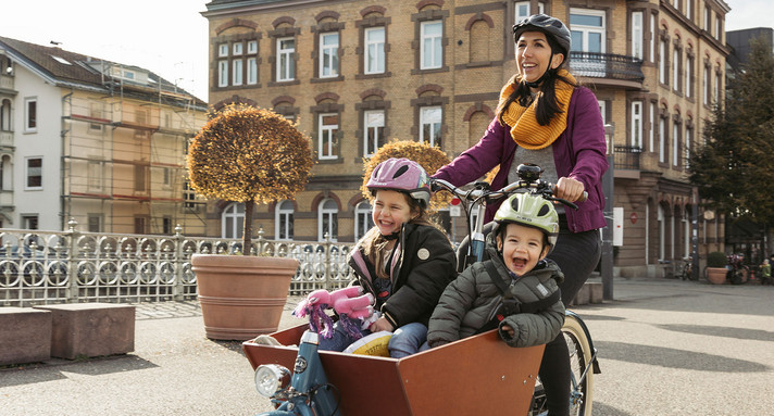 Zwei lachende Kinder sitzen in einem Lastenfahrrad und werden von einer Frau gefahren.